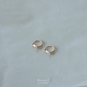 Puede incluir: Dos pendientes de aro dorados, cada uno con una sola gema rectangular transparente. Los pendientes se muestran sobre una superficie clara. La marca "Winnies & ATOWN JEWELRY" es visible en el centro inferior.