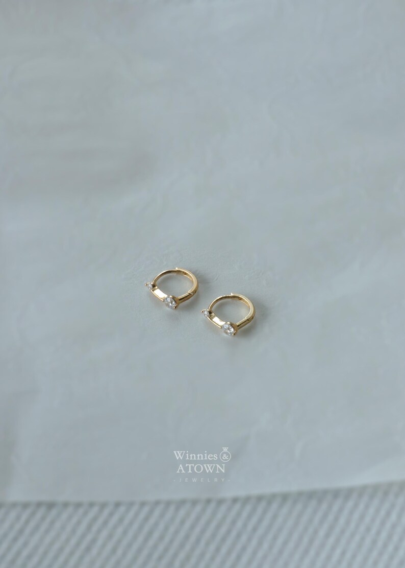 Puede incluir: Dos pendientes de aro dorados, cada uno con una sola gema rectangular transparente. Los pendientes se muestran sobre una superficie clara. La marca "Winnies & ATOWN JEWELRY" es visible en el centro inferior.