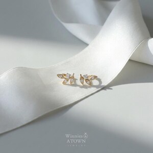 May include: A pair of gold-colored leaf-shaped earrings with small, clear gemstones. The earrings are displayed on a white satin ribbon. The brand name "Winnies & ATOWN JEWELRY" is visible in the lower right corner.