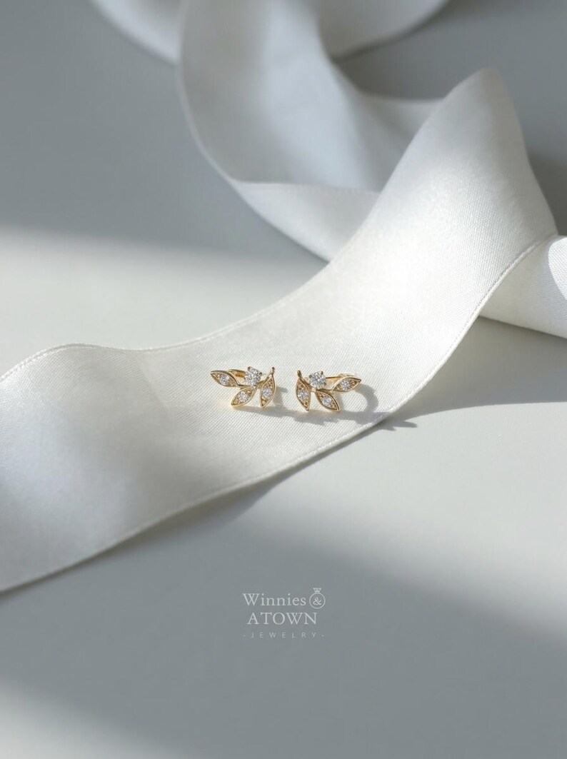 May include: A pair of gold-colored leaf-shaped earrings with small, clear gemstones. The earrings are displayed on a white satin ribbon. The brand name "Winnies & ATOWN JEWELRY" is visible in the lower right corner.