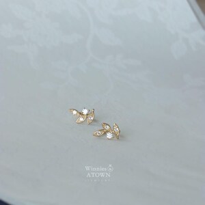 May include: A pair of gold-colored leaf-shaped stud earrings with clear, sparkling gemstones. The earrings are displayed on a light-colored surface with a subtle floral pattern. The brand name "Winnies ATOWN" is visible.