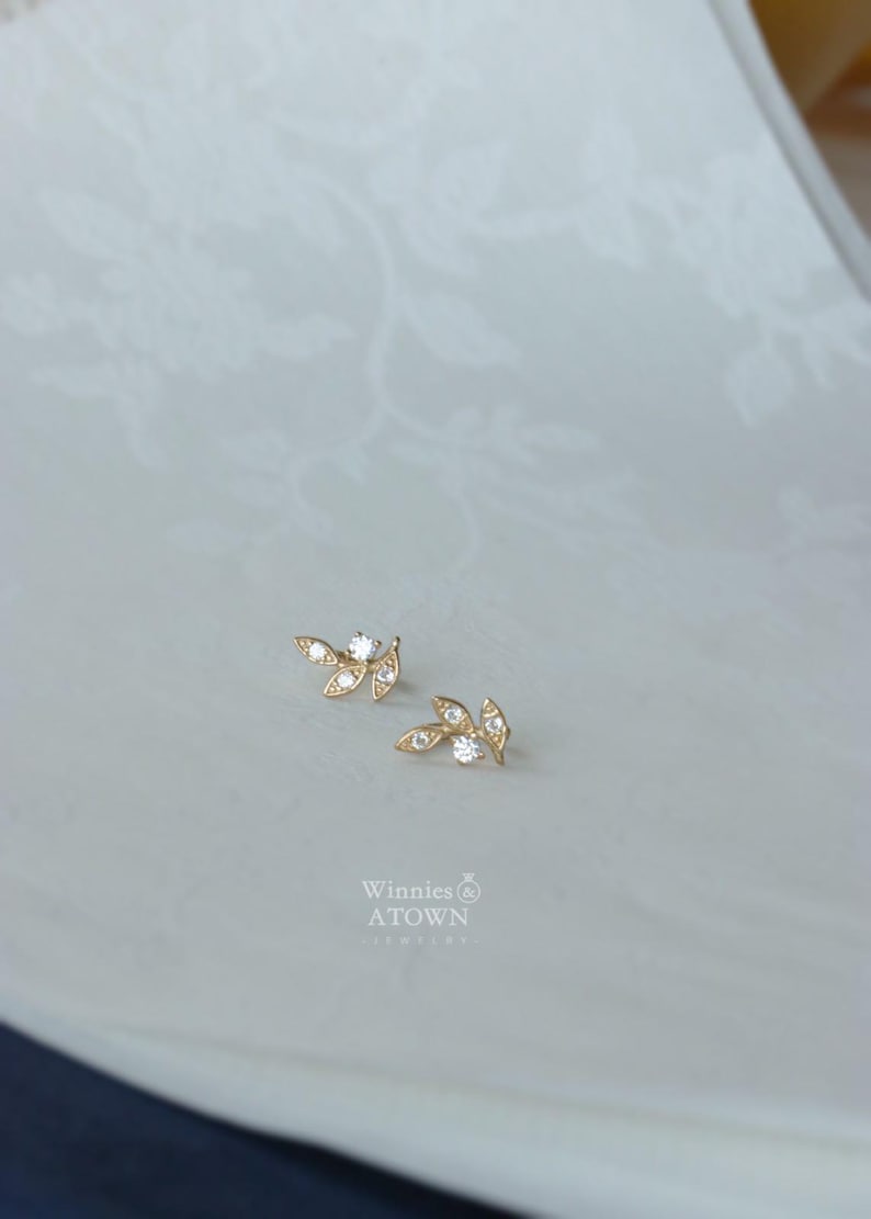 May include: A pair of gold-colored leaf-shaped stud earrings with clear, sparkling gemstones. The earrings are displayed on a light-colored surface with a subtle floral pattern. The brand name "Winnies ATOWN" is visible.