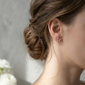 May include: A close-up of a person's ear adorned with a delicate gold earring. The earring features a floral design with small, sparkling gemstones. The person's hair is styled in a bun, and white flowers are visible in the background.