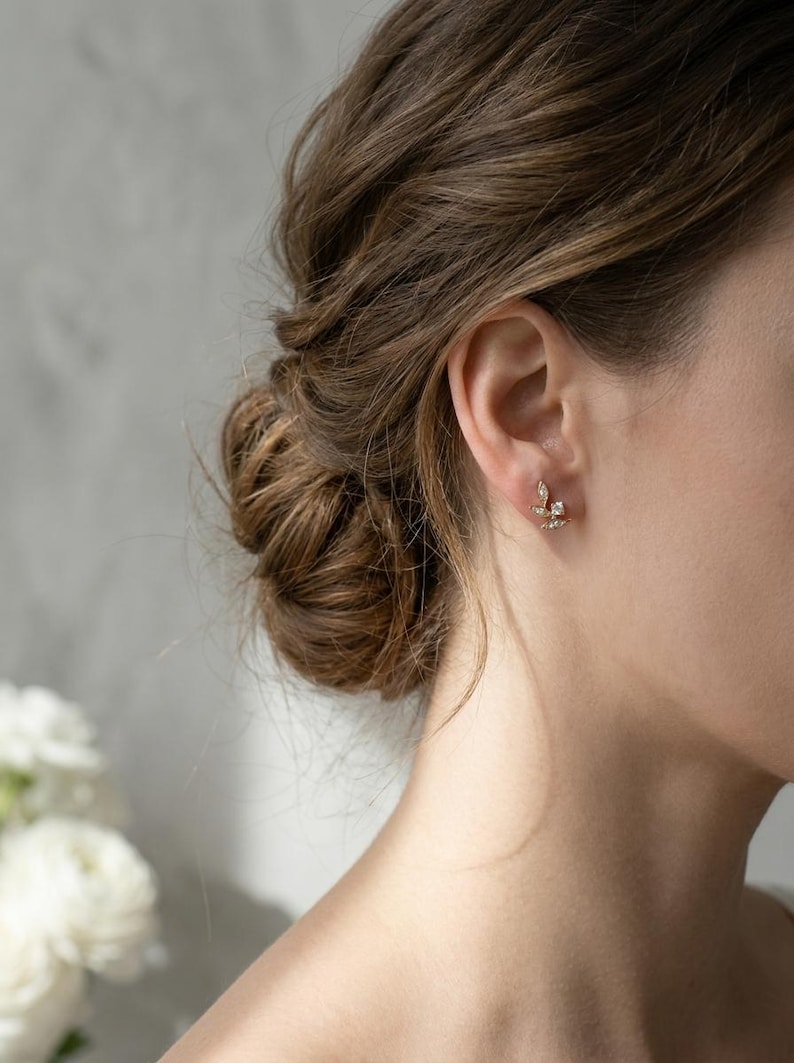 May include: A close-up of a person's ear adorned with a delicate gold earring. The earring features a floral design with small, sparkling gemstones. The person's hair is styled in a bun, and white flowers are visible in the background.