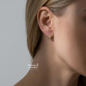May include: A close-up shot of a gold earring with a leaf design, adorned with small, sparkling gemstones. The earring is worn on the ear of a person with blonde hair. The jewelry is from Winnies & A Town Jewelry.