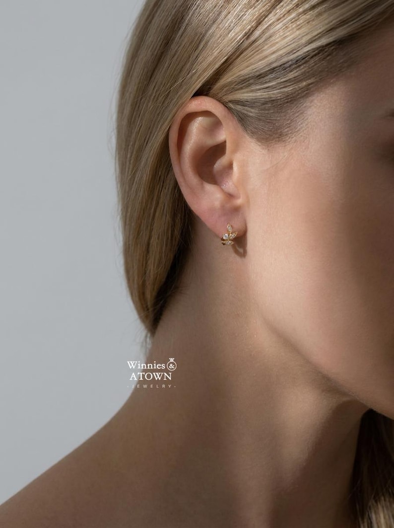 May include: A close-up shot of a gold earring with a leaf design, adorned with small, sparkling gemstones. The earring is worn on the ear of a person with blonde hair. The jewelry is from Winnies & A Town Jewelry.