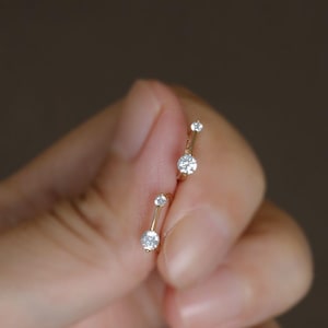Puede incluir: Un par de pendientes dorados con tres piedras preciosas redondas y transparentes. Los pendientes se sostienen en una mano sobre un fondo borroso. Los pendientes son de la marca "Winnies ATOWN Jewelry."