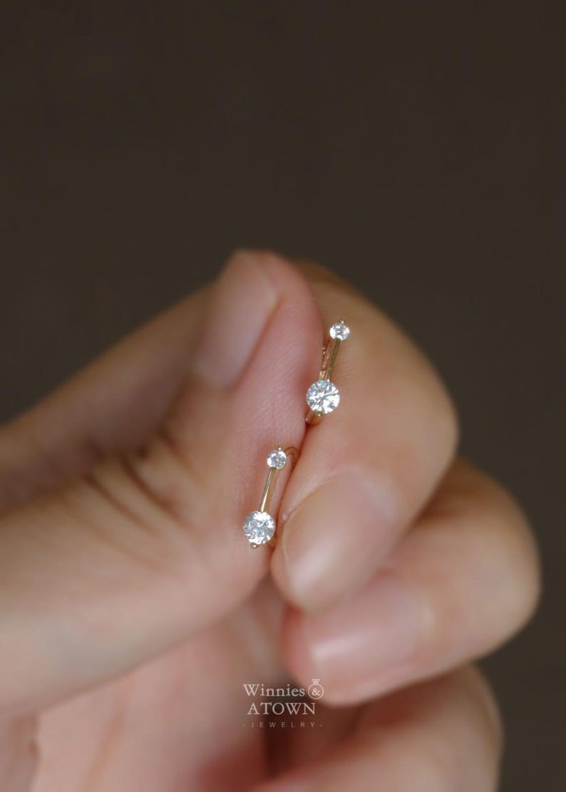 Puede incluir: Un par de pendientes dorados con tres piedras preciosas redondas y transparentes. Los pendientes se sostienen en una mano sobre un fondo borroso. Los pendientes son de la marca "Winnies ATOWN Jewelry."