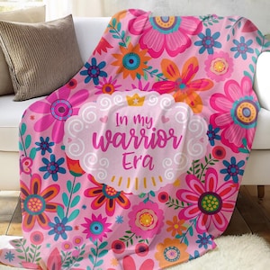 K&ouml;nnte beinhalten: Eine rosa Blumendecke mit dem Text "In my warrior Era" liegt &uuml;ber einem wei&szlig;en Sofa. Die Decke zeigt ein Muster aus gro&szlig;en rosa, orangefarbenen und blauen Blumen. Der Raum hat eine neutrale Farbpalette mit dekorativen Pflanzen.