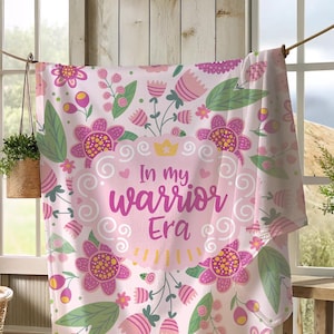K&ouml;nnte beinhalten: Eine rosa Blumendecke mit dem Text "In my warrior Era" h&auml;ngt an einer W&auml;scheleine. Die Decke hat ein sich wiederholendes Muster aus rosa und lila Blumen, gr&uuml;nen Bl&auml;ttern und gelben Akzenten. Der Hintergrund ist hellrosa.
