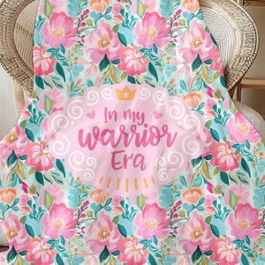 May include: A floral blanket with a light blue background and pink flowers. The blanket has the text "In my Warrior Era" in a pink oval with a crown. The blanket is draped over a wicker chair.