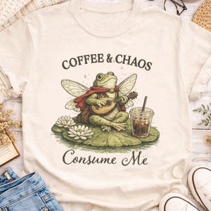 May include: Cream-colored t-shirt with the text "Coffee & Chaos Consume Me." The graphic features a frog playing a lute, wearing a cape, and holding a coffee drink. The frog is sitting on a lily pad.