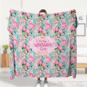 K&ouml;nnte beinhalten: Eine florale Decke mit hellblauem Hintergrund und einem Muster aus rosa und orangefarbenen Blumen mit gr&uuml;nen Bl&auml;ttern. Die Decke tr&auml;gt den Text "In my warrior Era" in einem rosa Oval.