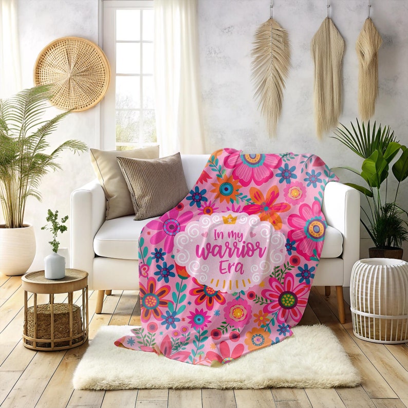 K&ouml;nnte beinhalten: Eine rosa Blumendecke mit dem Text "In my warrior Era" liegt &uuml;ber einem wei&szlig;en Sofa. Die Decke zeigt ein Muster aus gro&szlig;en rosa, orangefarbenen und blauen Blumen. Der Raum hat eine neutrale Farbpalette mit dekorativen Pflanzen.