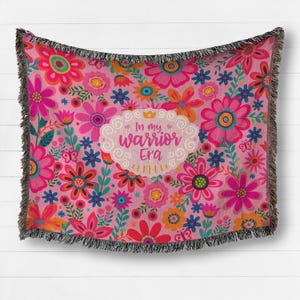 K&ouml;nnte beinhalten: Rosa Decke mit Blumenmuster und schwarzem Fransen. Die Decke zeigt eine Vielzahl bunter Blumen und den Text "In my warrior Era" in einem wei&szlig;en, wolkenf&ouml;rmigen Design.