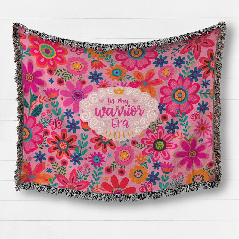 K&ouml;nnte beinhalten: Rosa Decke mit Blumenmuster und schwarzem Fransen. Die Decke zeigt eine Vielzahl bunter Blumen und den Text "In my warrior Era" in einem wei&szlig;en, wolkenf&ouml;rmigen Design.