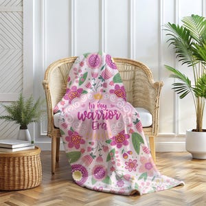 K&ouml;nnte beinhalten: Eine rosa-wei&szlig;e Blumen-Decke, die &uuml;ber einem Korbstuhl drapiert ist. Die Decke zeigt ein sich wiederholendes Muster aus rosa und lila Blumen, gr&uuml;nen Bl&auml;ttern und dem Text "In my warrior Era". Der Stuhl steht vor einer wei&szlig;en Wand mit einer Pflanze im Hintergrund.
