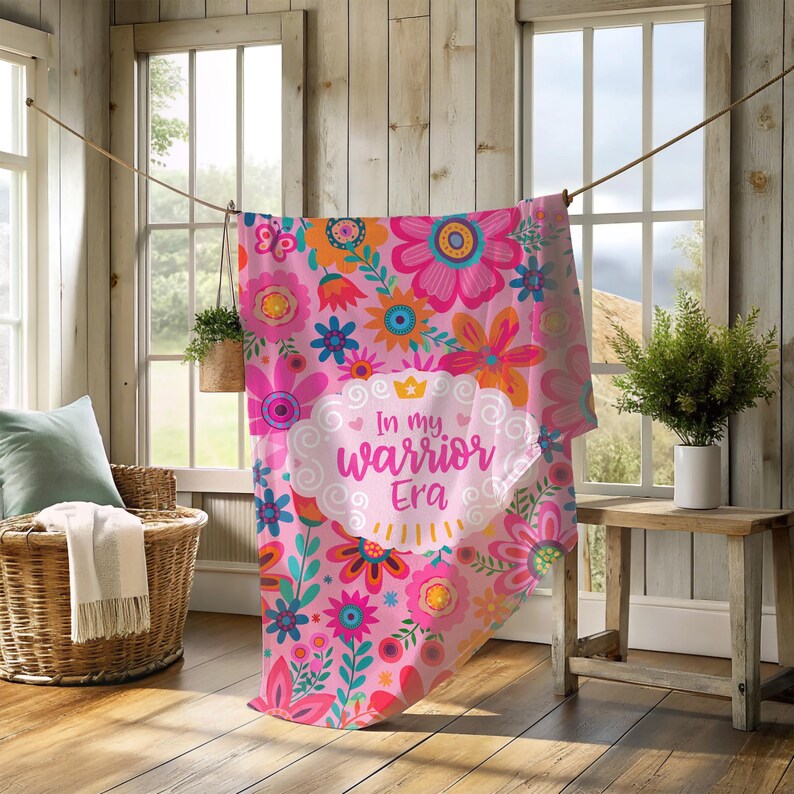 K&ouml;nnte beinhalten: Eine rosa Blumendecke mit einer Vielzahl farbenfroher Blumen, darunter rosa, orange und blau. Die Decke hat den Text "In my warrior Era" in einem wei&szlig;en wolkenf&ouml;rmigen Design. Die Decke h&auml;ngt an einer W&auml;scheleine.