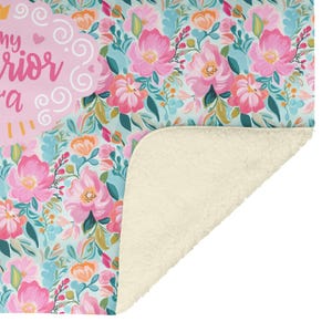 K&ouml;nnte beinhalten: Eine floral gemusterte Decke mit hellblauem Hintergrund und rosa Blumen. Die Decke hat ein weiches, cremefarbenes Sherpa-Futter. Der Text "my warrior" ist auf der Decke zu sehen.