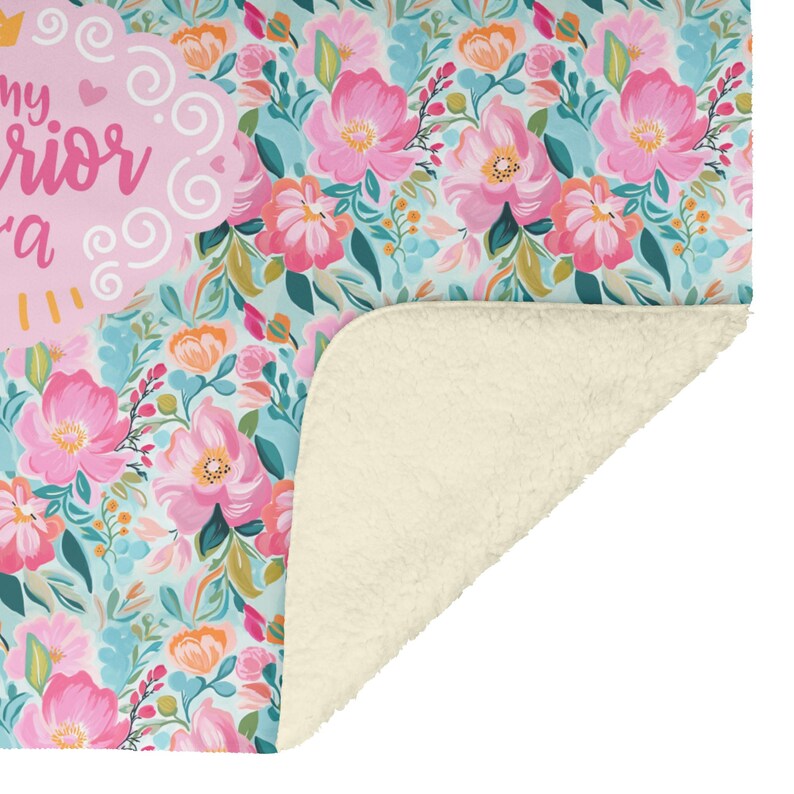 K&ouml;nnte beinhalten: Eine floral gemusterte Decke mit hellblauem Hintergrund und rosa Blumen. Die Decke hat ein weiches, cremefarbenes Sherpa-Futter. Der Text "my warrior" ist auf der Decke zu sehen.