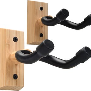 May include: Two guitar wall mounts. Each mount features a black, curved cradle attached to a light-colored, rectangular wooden base. The bases are mounted with black screws. The mounts are designed to hold a guitar.