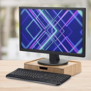 Może przedstawiać: Czarny monitor komputera wyświetlający niebiesko-fioletowy geometryczny wz&oacute;r, spoczywający na bambusowej podstawce pod monitor z szufladą. Czarna klawiatura znajduje się przed monitorem i podstawką. Zestaw znajduje się na jasnej drewnianej powierzchni.