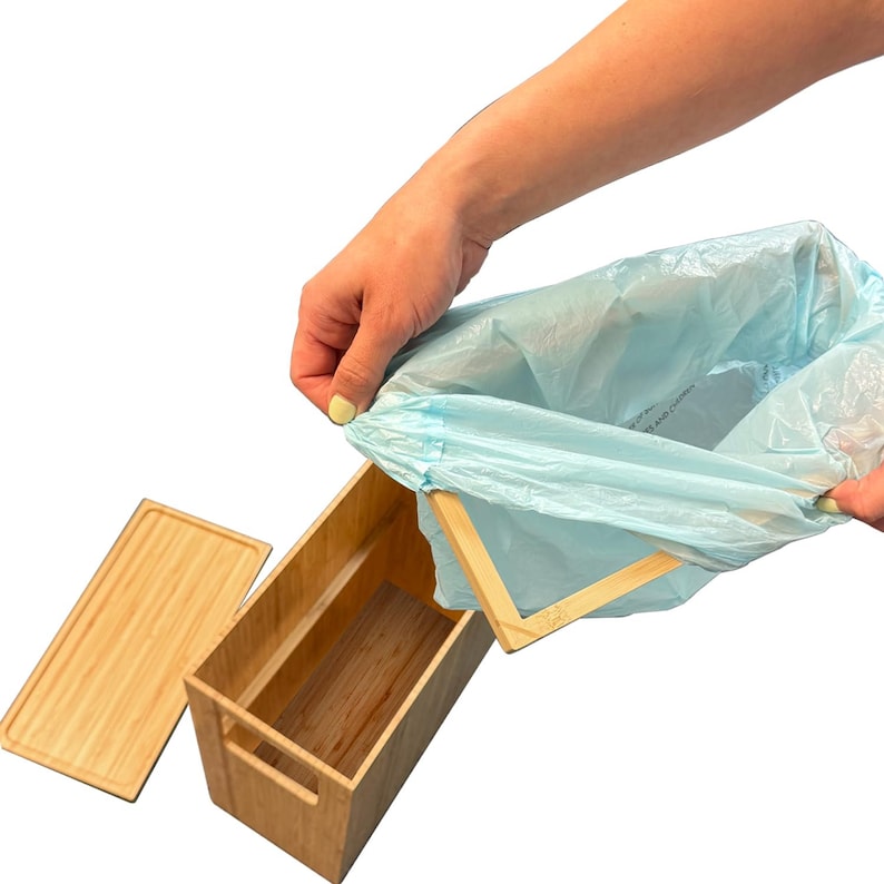May include: A light blue plastic bag is being placed into a wooden box with a lid. The box has a handle on the side. The box is made of bamboo.
