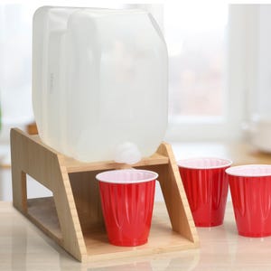 Op de afbeelding: Een heldere, rechthoekige waterkan met een kraan staat op een bamboe standaard. Drie rode plastic bekers met witte randen staan ervoor. De set is voor het gemakkelijk afgeven van water.