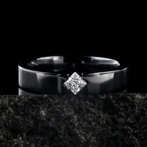 Può includere: Un anello nero con un diamante a taglio quadrato come pietra centrale. L'anello ha una fascia piatta e larga e una finitura lucida. Il diamante è incastonato in una lunetta.