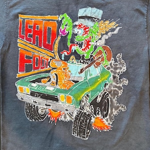 May include: Dark gray t-shirt with a colorful graphic. The graphic features the text "LEAD FOOT" in orange and red, a green car with a cartoon character playing a guitar, and flames. The car has large tires and is emitting smoke.
