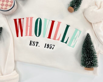 May include: White sweatshirt with a red, pink, green, and blue embroidered arch design that reads "WHOOVILLE EST. 1957". Two small green artificial Christmas trees are on the sweatshirt.