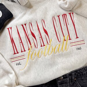 Op de afbeelding: Een lichtgrijs sweatshirt met een rood en goud geborduurd logo met de tekst "Kansas City Football est. 1963".