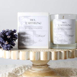 Puede incluir: Un tarro de vela blanco con una etiqueta negra que dice "HEY. EARTHLING SOOTHING SWEET JASMINE & EVOCATIVE PATCHOULI HAND POURED NATURAL SOY WAX CANDLE made with love" y una caja blanca con la misma etiqueta. La vela y la caja están sobre un soporte para pasteles de madera blanco y marrón. Un ramo de lavanda seca está en el lado izquierdo del soporte.