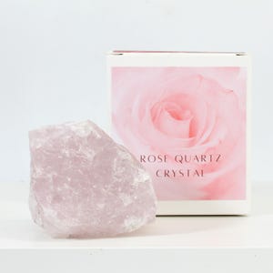 Könnte beinhalten: Ein großer, hellrosa Rosenquarzkristall mit einer rauen, natürlichen Textur. Der Kristall liegt auf einer weißen Oberfläche neben einer weißen Schachtel mit einer rosa Rose und dem Text "ROSE QUARTZ CRYSTAL".
