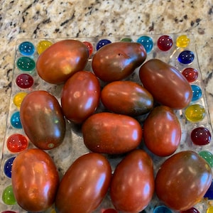 May include: A collection of 12 dark brown heirloom tomatoes on a glass plate with colorful glass accents. The tomatoes are oval shaped and have a glossy finish.