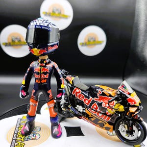 Könnte beinhalten: Figur einer Rennfahrerin in einem schwarzen und orangefarbenen Red Bull Rennanzug, neben einem passenden Motorrad. Das Motorrad und der Helm haben das Red Bull Logo. Der Helm hat den Text "GIVES YOU WINGS".