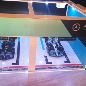 K&ouml;nnte beinhalten: Ein Miniaturmodell einer Formel-1-Garage mit zwei Rennwagen im Inneren. Die Autos sind beide schwarz und silber mit der Nummer 44 und dem Namen "Lewis Hamilton" darauf. Die Garage ist aus Holz und hat ein blaues und gr&uuml;nes Interieur. Die Garage hat ein Schild mit der Aufschrift "AMG PETRONAS Formula One Team".
