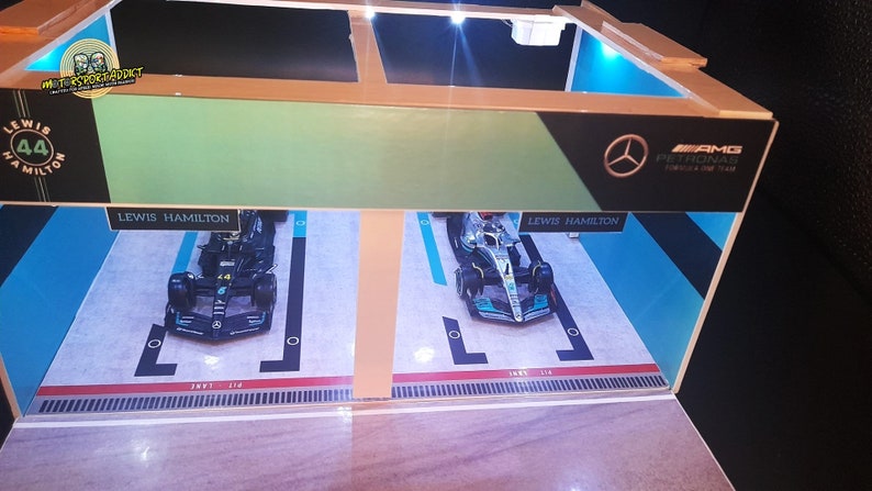 K&ouml;nnte beinhalten: Ein Miniaturmodell einer Formel-1-Garage mit zwei Rennwagen im Inneren. Die Autos sind beide schwarz und silber mit der Nummer 44 und dem Namen "Lewis Hamilton" darauf. Die Garage ist aus Holz und hat ein blaues und gr&uuml;nes Interieur. Die Garage hat ein Schild mit der Aufschrift "AMG PETRONAS Formula One Team".