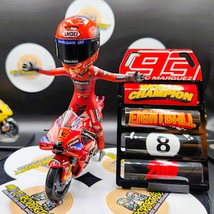Peut inclure: Figurine de moto Ducati rouge avec un pilote portant un casque et une combinaison rouges. La figurine est à côté d'un présentoir noir avec le numéro 93 et les mots "World Champion". D'autres motos jouets sont en arrière-plan.