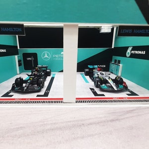 K&ouml;nnte beinhalten: Ein Miniaturmodell einer Formel-1-Boxengasse mit zwei Rennwagen, einem schwarzen und einem silbernen, beide mit Petronas-Branding. Die Boxengasse ist in den Farben des Teams, gr&uuml;n und schwarz, und dem Namen des Fahrers, Lewis Hamilton, dekoriert.