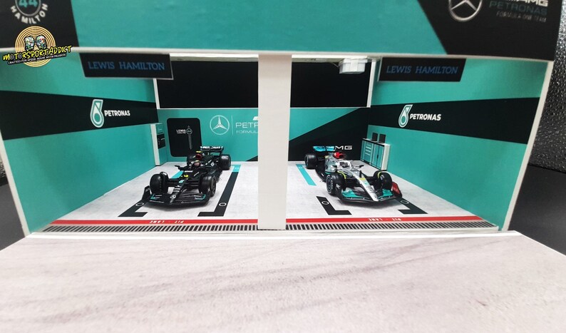 K&ouml;nnte beinhalten: Ein Miniaturmodell einer Formel-1-Boxengasse mit zwei Rennwagen, einem schwarzen und einem silbernen, beide mit Petronas-Branding. Die Boxengasse ist in den Farben des Teams, gr&uuml;n und schwarz, und dem Namen des Fahrers, Lewis Hamilton, dekoriert.