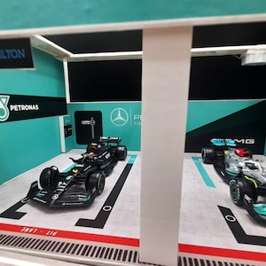 K&ouml;nnte beinhalten: Ein Modell einer Formel-1-Rennbox mit zwei Rennwagen im Inneren. Die Box ist in einem Teal- und Schwarz-Farbschema mit dem Petronas-Logo und den Worten "Lewis Hamilton" bemalt.