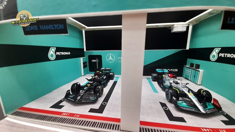 K&ouml;nnte beinhalten: Ein Modell einer Formel-1-Rennbox mit zwei Rennwagen im Inneren. Die Box ist in einem Teal- und Schwarz-Farbschema mit dem Petronas-Logo und den Worten "Lewis Hamilton" bemalt.