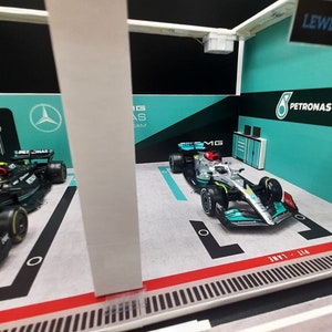 K&ouml;nnte beinhalten: Ein Miniaturmodell einer Formel-1-Rennwagen-Garage mit zwei Rennwagen, einem schwarzen und einem silbernen, mit den Logos von Petronas und Mercedes. Die Garage verf&uuml;gt &uuml;ber eine Boxengasse und ein Schild mit der Aufschrift "Lewis Hamilton".