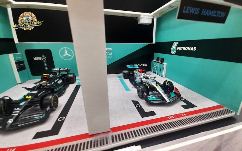 K&ouml;nnte beinhalten: Ein Miniaturmodell einer Formel-1-Rennwagen-Garage mit zwei Rennwagen, einem schwarzen und einem silbernen, mit den Logos von Petronas und Mercedes. Die Garage verf&uuml;gt &uuml;ber eine Boxengasse und ein Schild mit der Aufschrift "Lewis Hamilton".