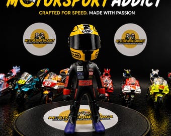 May include: A figurine of a motorcycle racer wearing a yellow and black helmet and a black racing suit with Honda Britain branding. The figure stands on a circular display base, surrounded by several miniature toy motorcycles on a black surface.