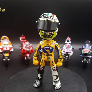 May include: A yellow and black figurine of a motorcycle racer wearing a helmet with the number 46 on the front. The figurine is standing in front of five miniature motorcycles.