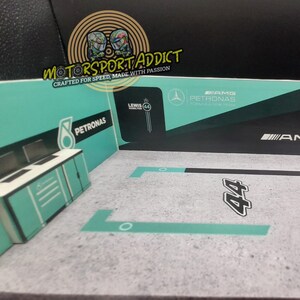 May include: A model of a Formula 1 race car garage with a green and black color scheme. The garage has a sign that says "PETRONAS" and "AMG". The garage also has a sign that says "Lewis Hamilton 44".