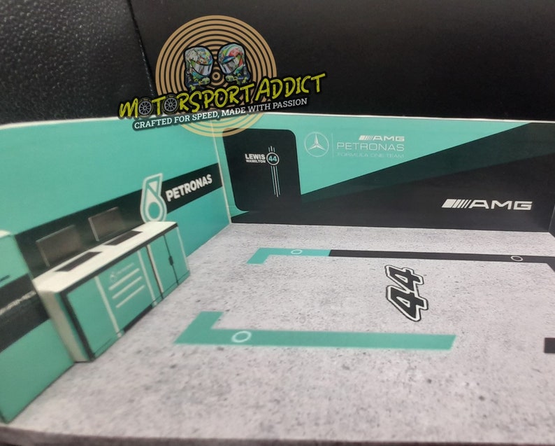 May include: A model of a Formula 1 race car garage with a green and black color scheme. The garage has a sign that says "PETRONAS" and "AMG". The garage also has a sign that says "Lewis Hamilton 44".