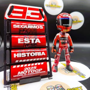 Könnte beinhalten: Ein rot-schwarzer Display-Ständer mit den Texten "SEGUIMOS, ESTA, HISTORIA" und "2025 MOTOGP WORLD CHAMPION". Eine Figur eines Rennfahrers in rotem Anzug und Helm steht daneben.